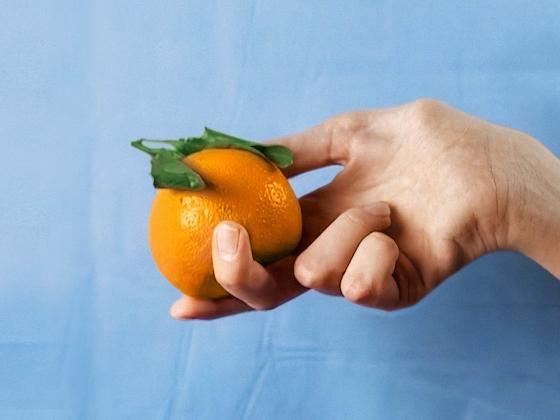 A woman's hand holds a bright orange against a light blue, slightly crumpled background of fabric.