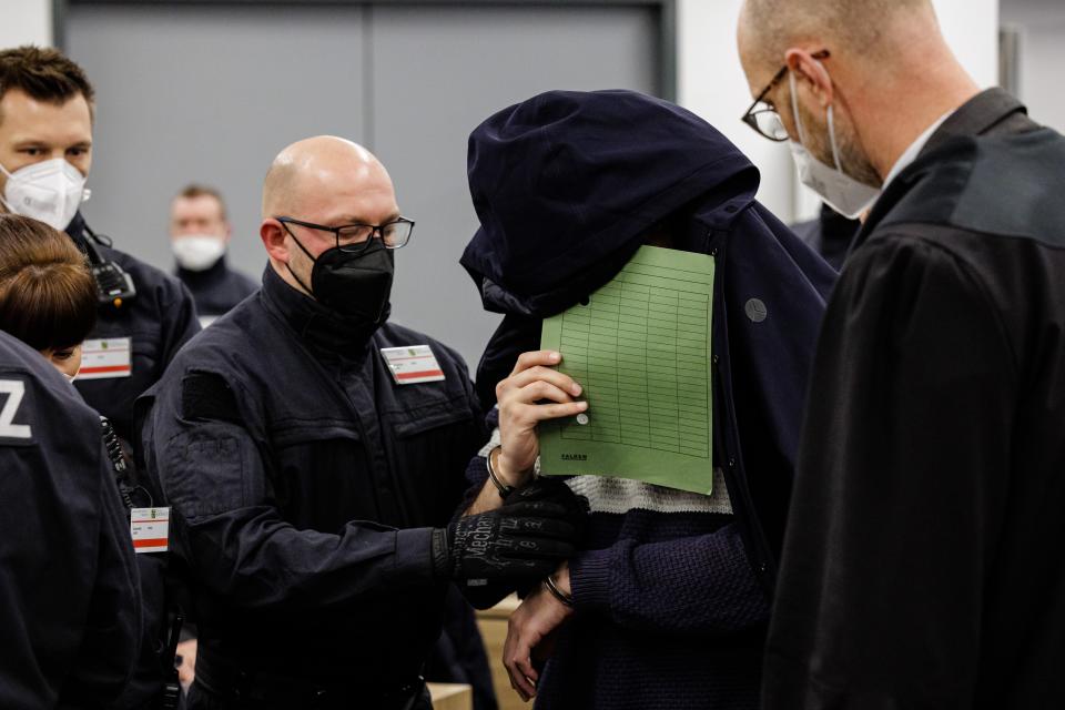 Ein Angeklagter (M) verdeckt sein Gesicht, während in einen Saal des Oberlandesgerichts Dresden geführt wird