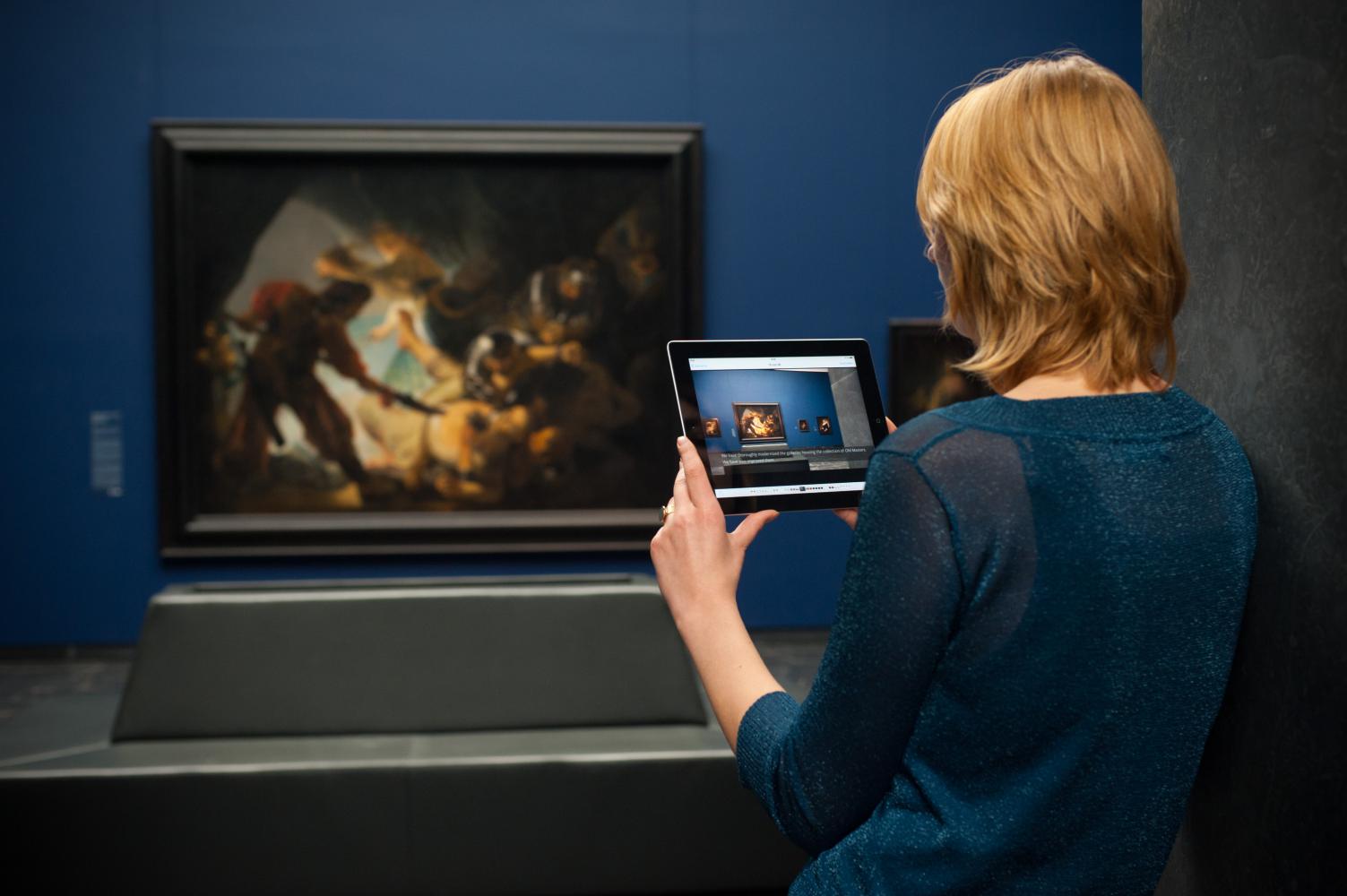  Eine Frau hält ein Tablet in den Händen vor einem Gemälde in einem Ausstellungsraum des Städel Museums