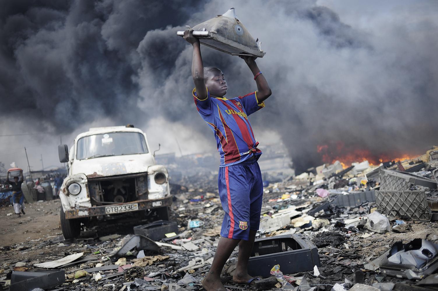 Kai Löffelbein, Afrika, Ghana, Accra, Agbogbloshie, 20.09.2011: Ein Junge wirft einen Fernseher mehrere Male auf den Boden, um an das Metall zu kommen. Im Hintergrund werden Reifen verbrannt