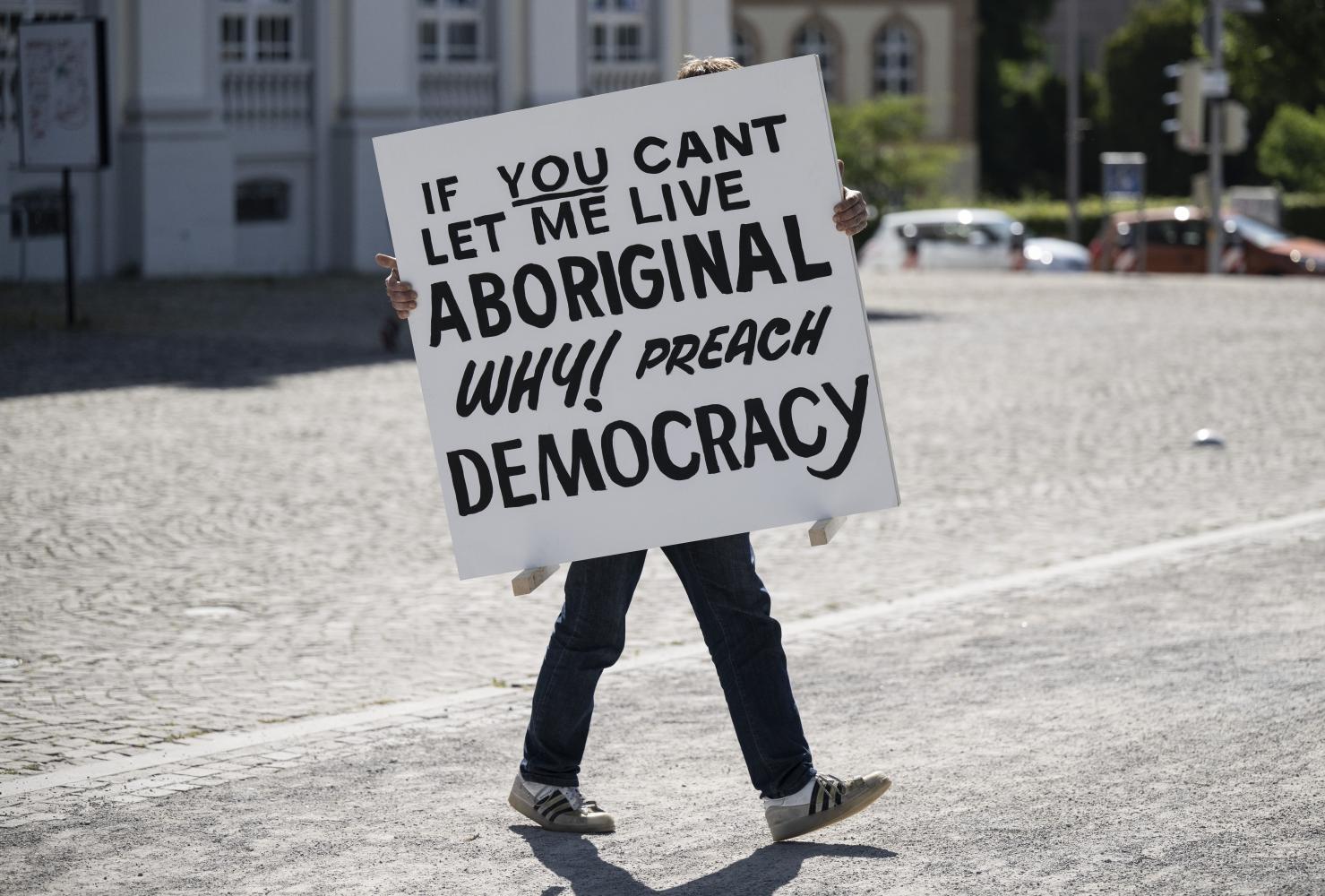 Ein Mann trägt ein Schild mit der Aufschrift "If you can’t let me live aboriginal, why preach democrazy" zu einer Installation, die sich mit dem Schicksal der Aborigines in Australien befasst