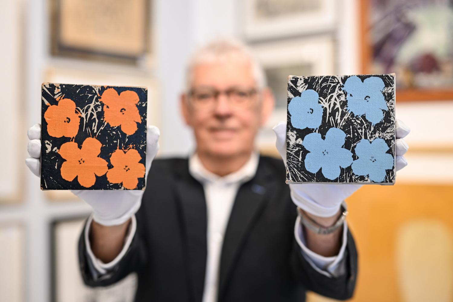 Udo Geble von Auktionshaus Geble hält zwei Warhol-Bilder mit dem Namen "Flowers" in den Händen